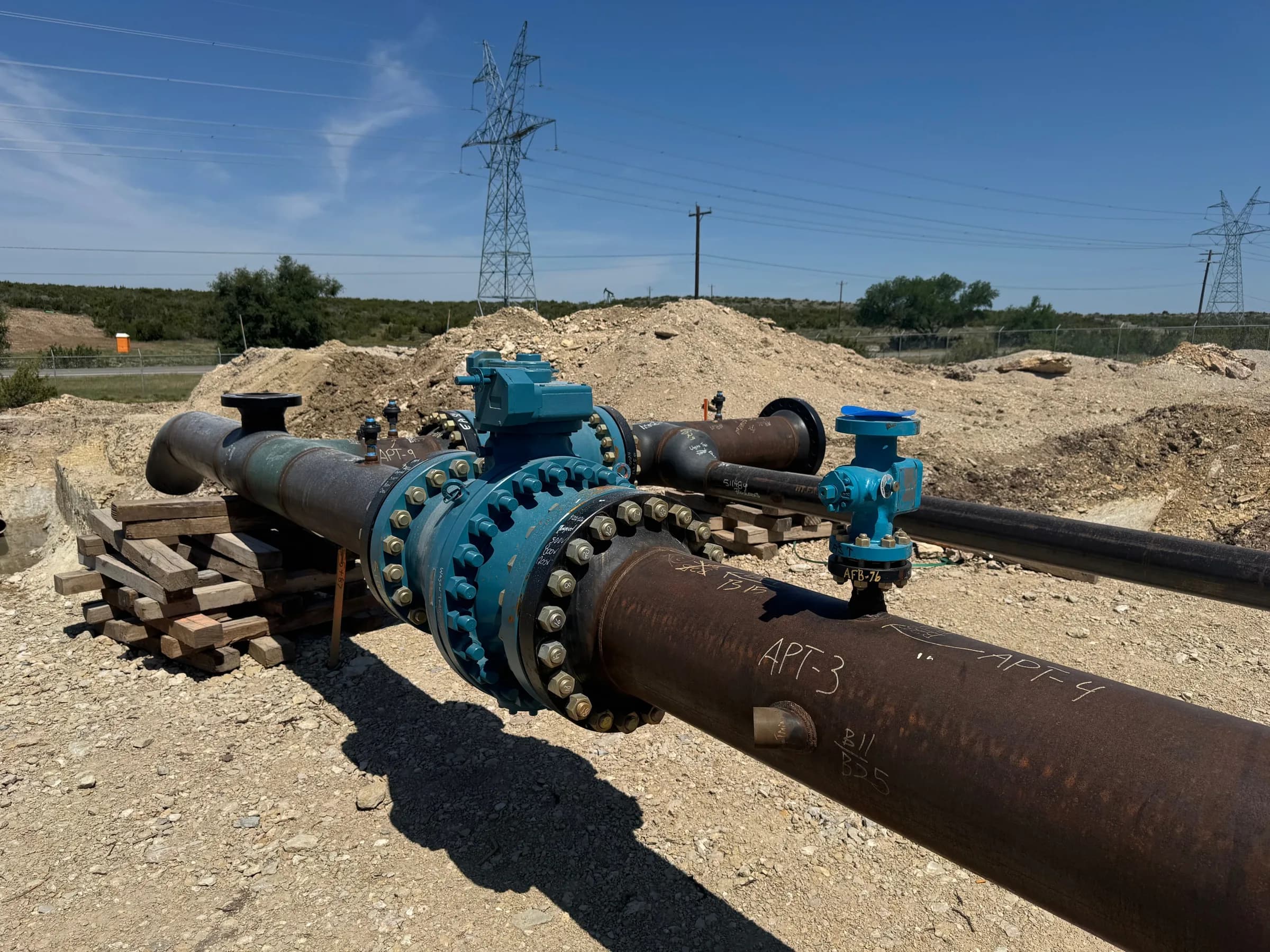 Flanged pipeline valve assembly with blue-painted fittings on a coated steel line at a graded worksite under a clear sky