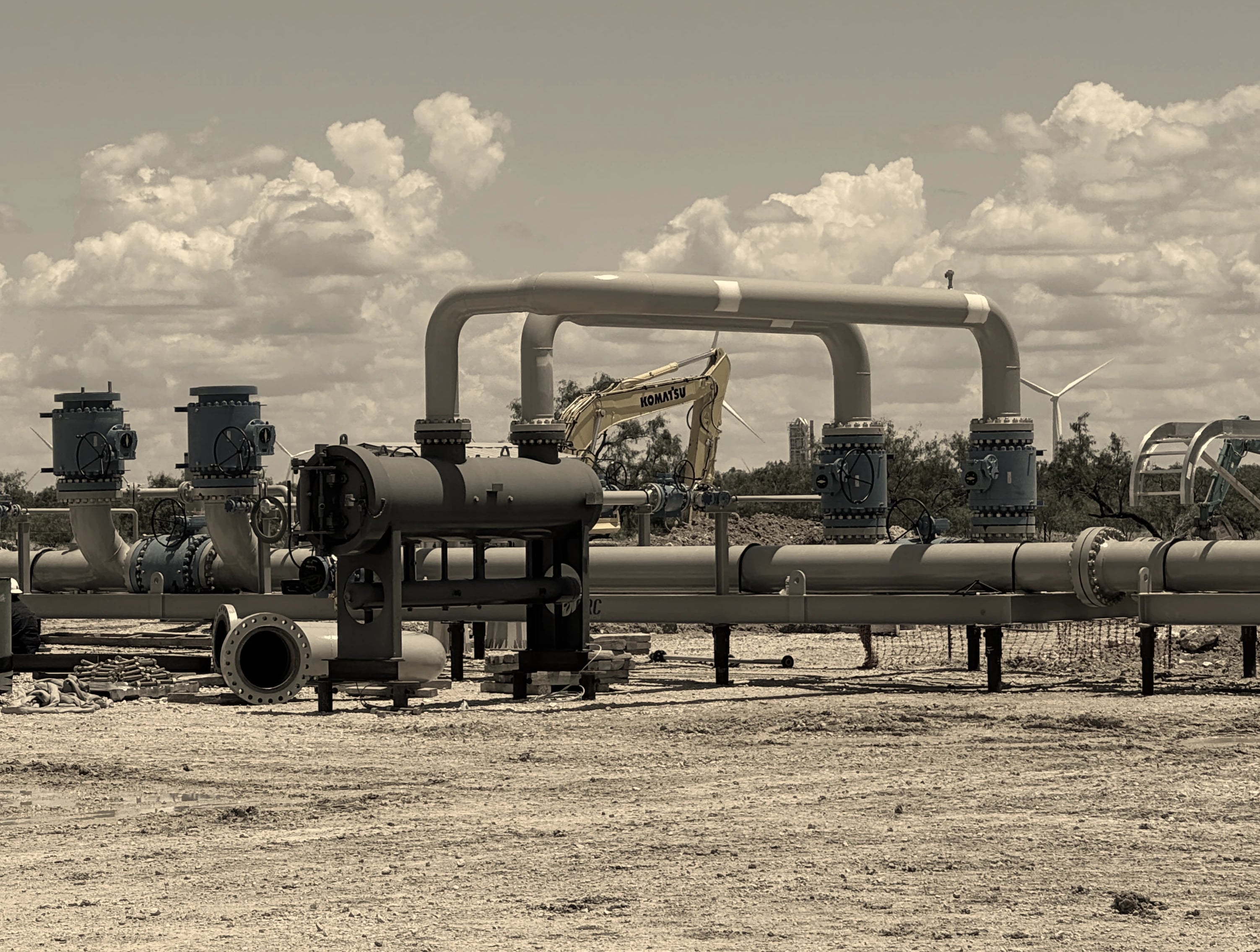 Aboveground pipeline manifold with arched piping and valve stacks at a construction site, an excavator and wind turbines behind